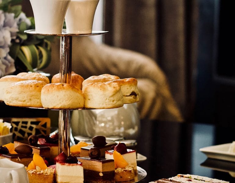 Silver afternoon tea stand with scones, cakes and desserts, on table with teacups, sandwiches and floral decor.