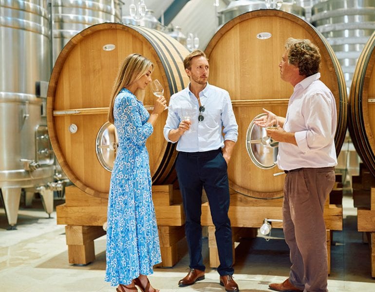 Three people enjoying a wine tasting in a winery, standing in front of large wooden barrels and stainless steel tanks.