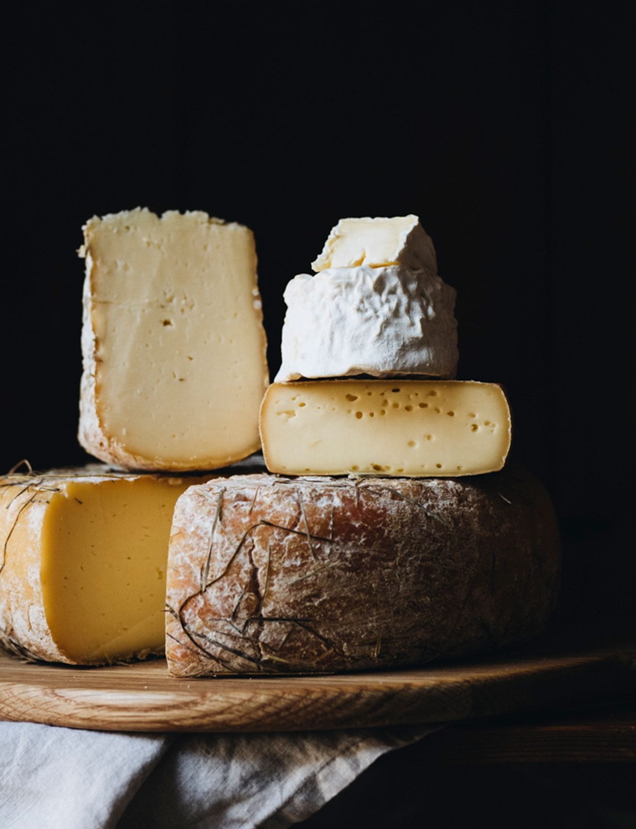 Assorted artisanal cheeses stacked on a wooden board against a dark background, with a cloth partially draped underneath.