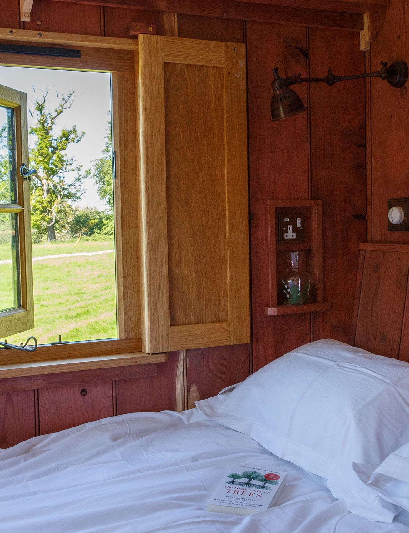 Wooden hut bedroom with white bedding, open window shutters and views of green fields outside.