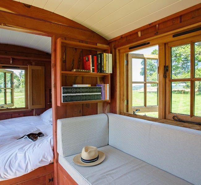 Wooden hut interior, cushioned window bench, bookshelves, large windows with field view and bedroom with bed in view.