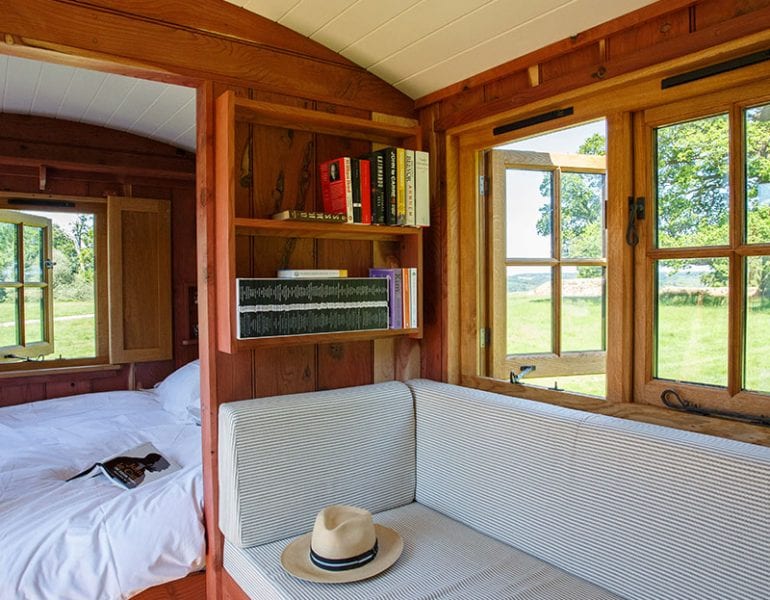 Wooden hut interior, cushioned window bench, bookshelves, large windows with field view and bedroom with bed in view.