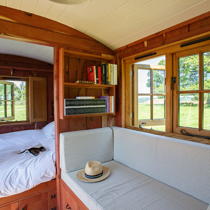 Wooden hut interior, cushioned window bench, bookshelves, large windows with field view and bedroom with bed in view.