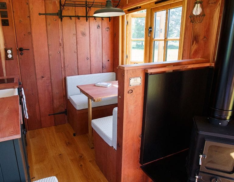 Interior of redwood drovers hut cabin with dining booth by window, lighting above table and black log burner.