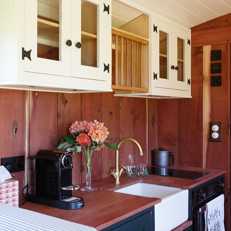 Compact kitchen, wood panelled walls, white cabinets, a farmhouse sink, coffee machine and vase of flowers on counter.