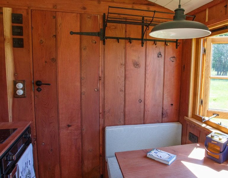 Compact wooden kitchen and dining area with natural timber walls, bench seating, table, stove and window letting natural light.