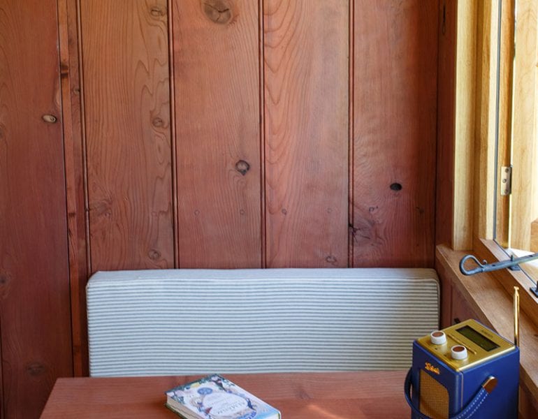 Close-up of small wooden dining table with book and radio, timber-clad walls and cushioned bench beside open window.