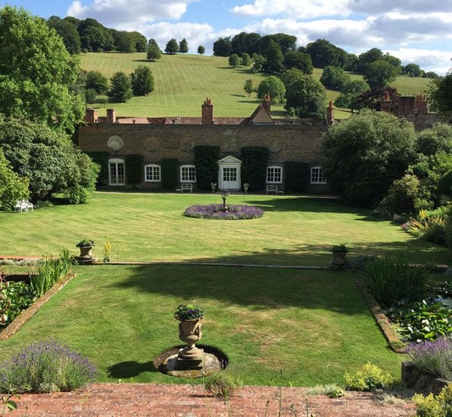 Formal garden with manicured lawn, central urn and flowerbeds, leading to an ivy-clad brick house backed by rolling green hills.