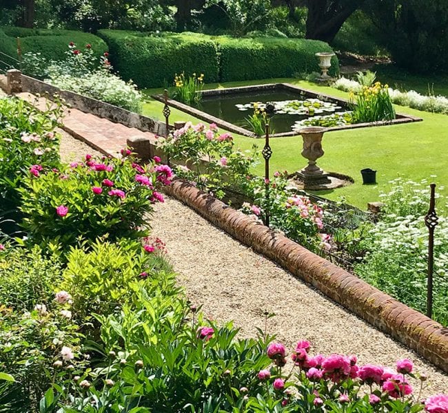 Gravel path bordered by blooming pink flowers, leading to lawn with square lily pond and stone urns in manicured garden.