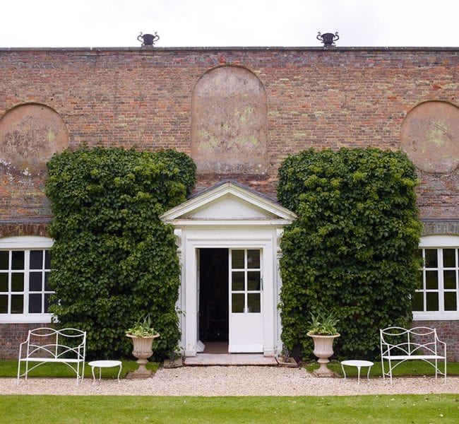 Formal garden with manicured lawn, central urn and flowerbeds, leading to an ivy-clad brick house backed by rolling green hills.