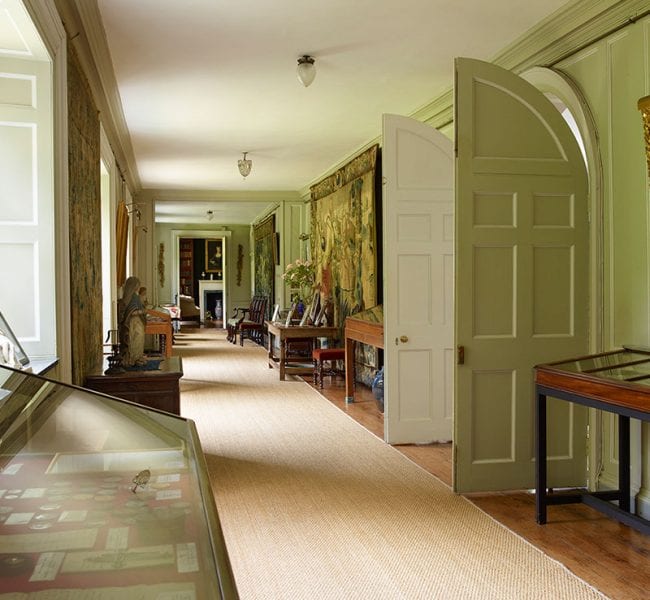 Formal garden with manicured lawn, central urn and flowerbeds, leading to an ivy-clad brick house backed by rolling green hills.