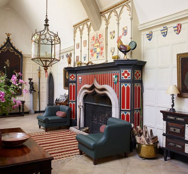 Formal garden with manicured lawn, central urn and flowerbeds, leading to an ivy-clad brick house backed by rolling green hills.