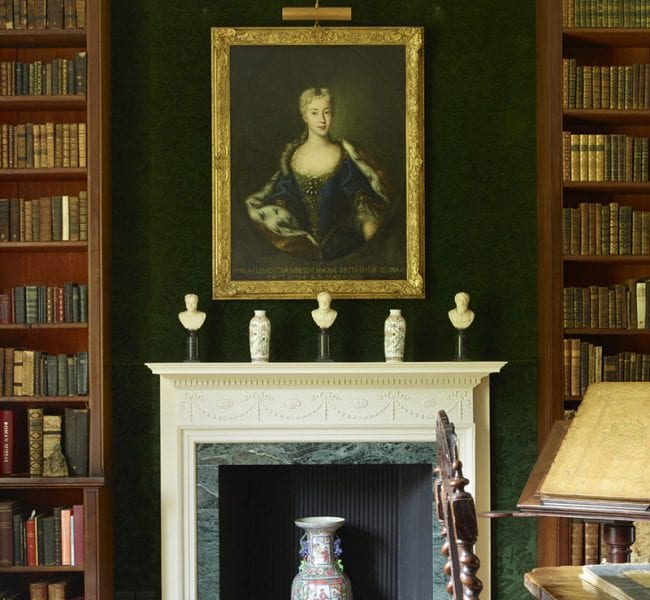 Formal garden with manicured lawn, central urn and flowerbeds, leading to an ivy-clad brick house backed by rolling green hills.