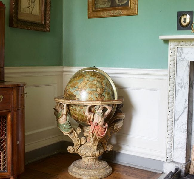 Formal garden with manicured lawn, central urn and flowerbeds, leading to an ivy-clad brick house backed by rolling green hills.