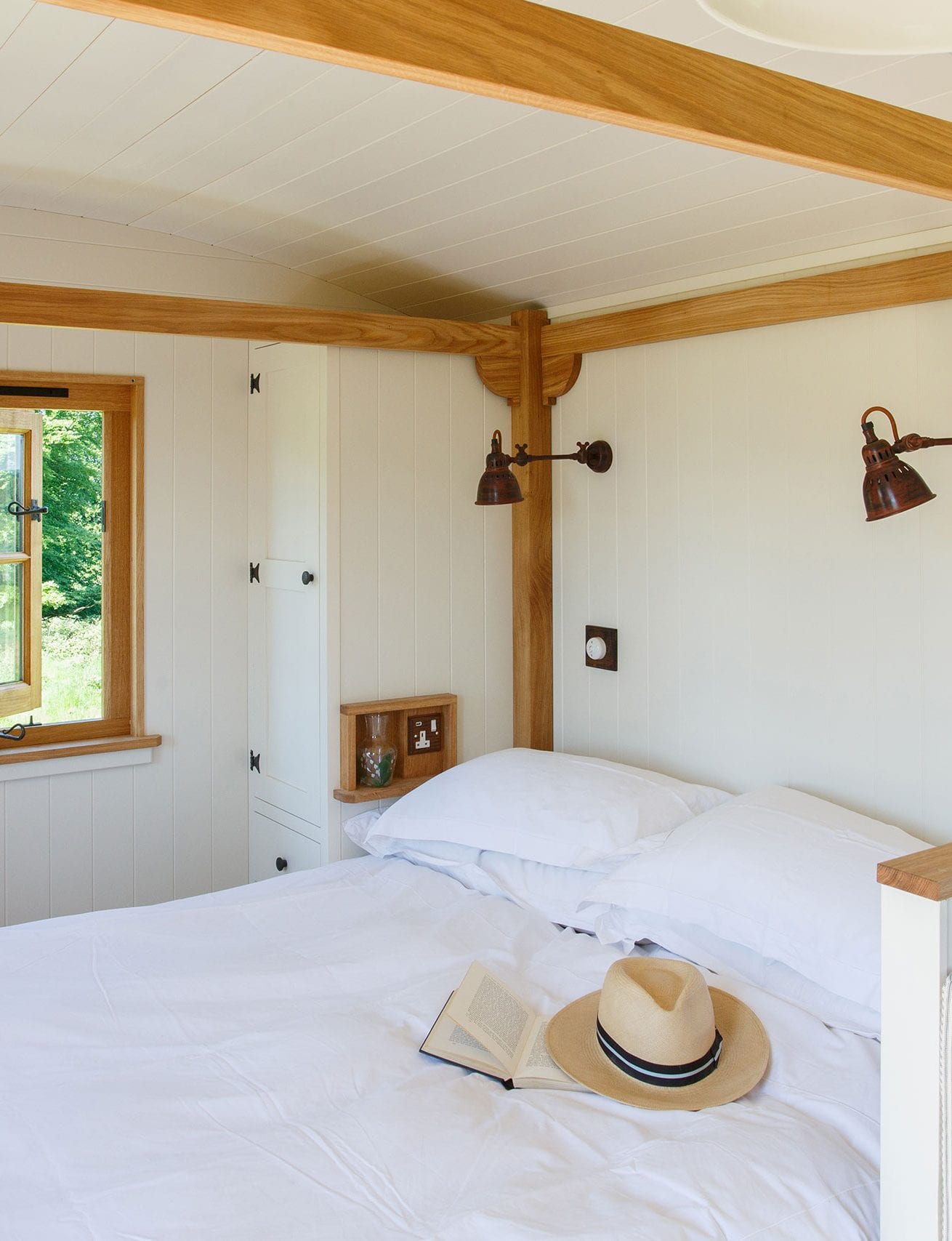 Bedroom with white walls, oak wood beams, king-size bed, open window, white bedding and straw hat resting on duvet.