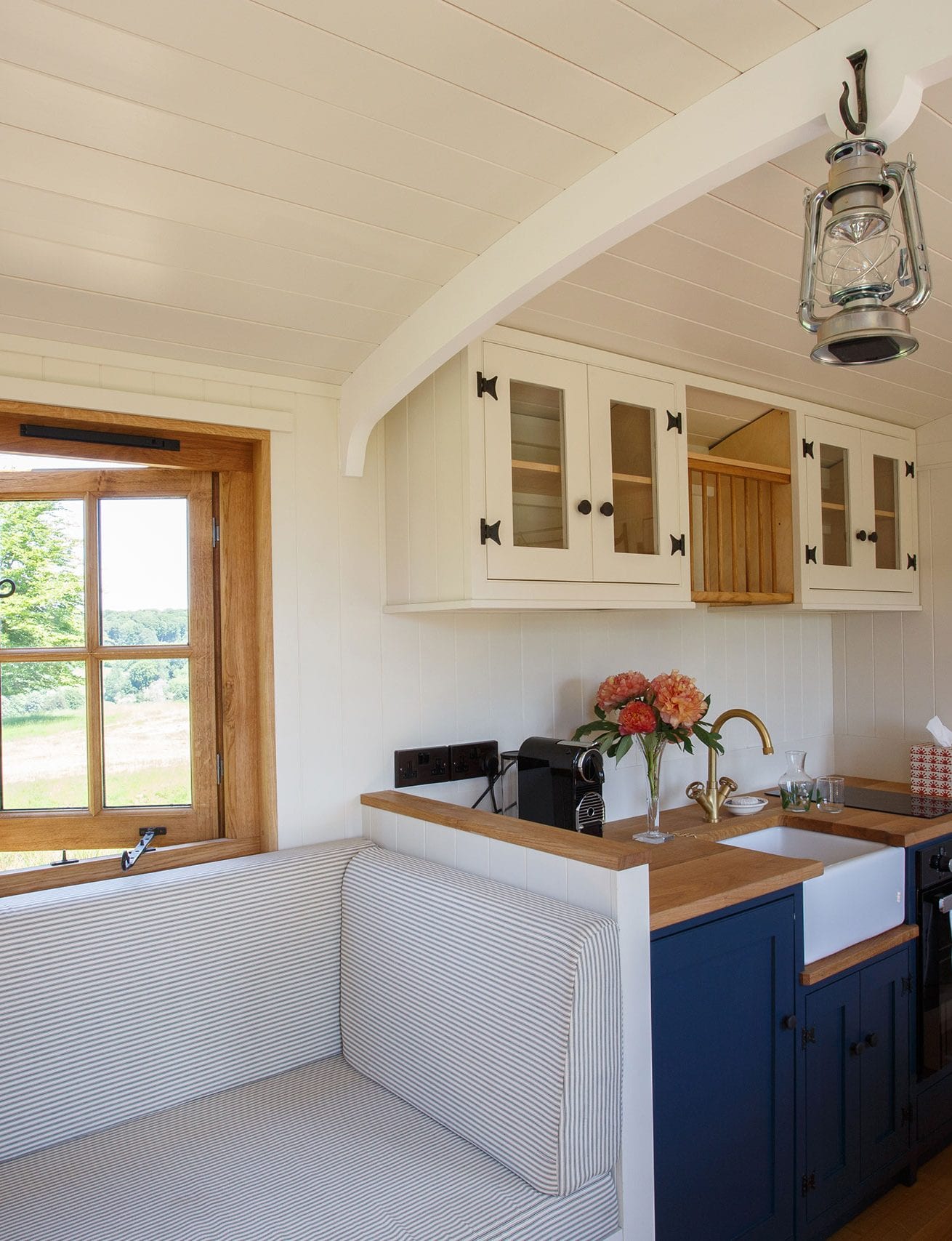 Drovers' hut kitchen with navy cabinets, white sink, cushioned bench, wooden windows and hanging lantern light.