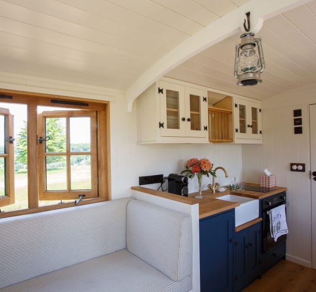 Drovers' hut kitchen with navy cabinets, white sink, cushioned bench, wooden windows and hanging lantern light.