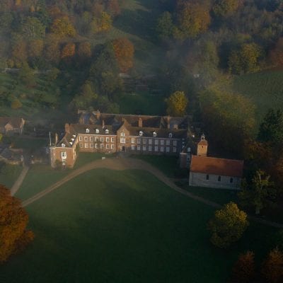 Aerial view of a large country house with autumn trees, mist and long shadows across the surrounding lawns and hills.