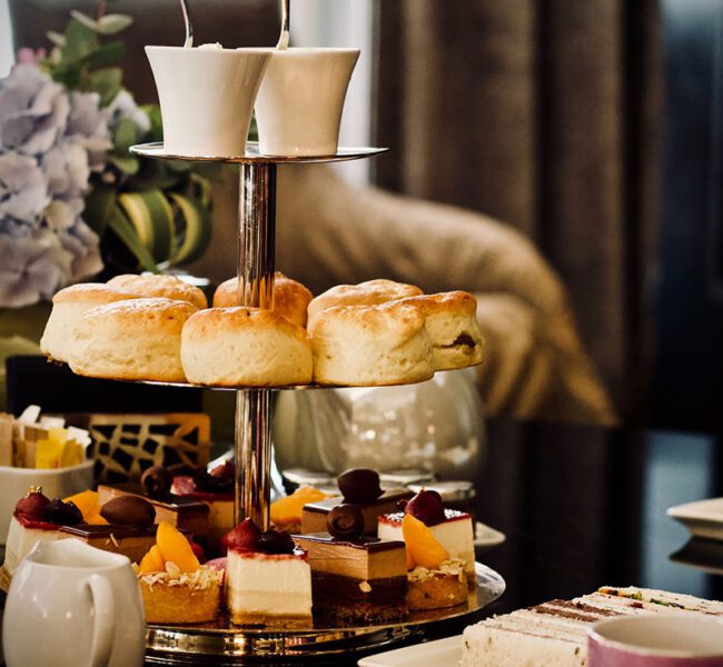 Elegant afternoon tea stand with scones, cakes and desserts, set on a table with teacups and floral decor.