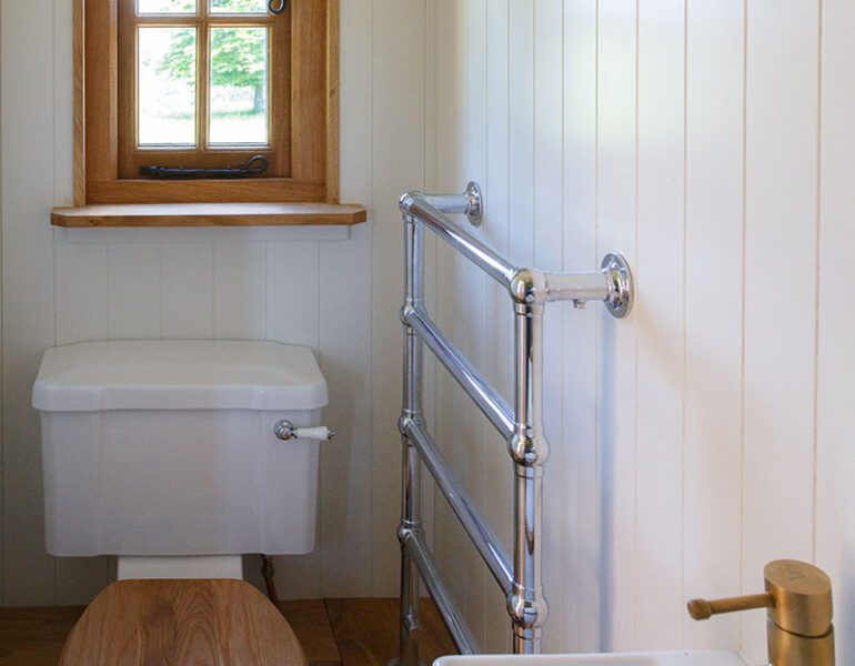 Compact bathroom - white panelled walls, toilet with wooden seat, chrome towel rail, sink, brass tap, window with wooden frame.