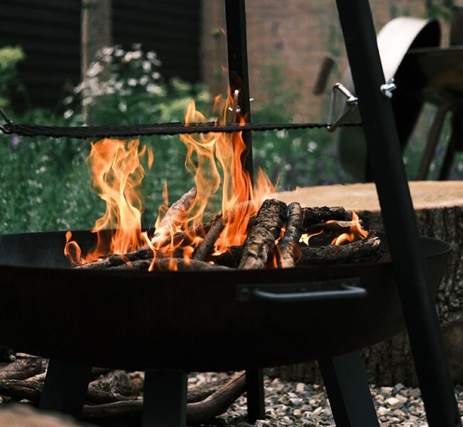Lit fire pit with flames rising from burning logs, surrounded by rustic wooden seating in an outdoor garden setting.