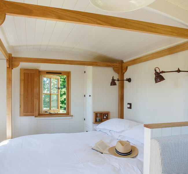 Bedroom with white walls, oak wood beams, a double bed, open window and a straw hat resting on the duvet.