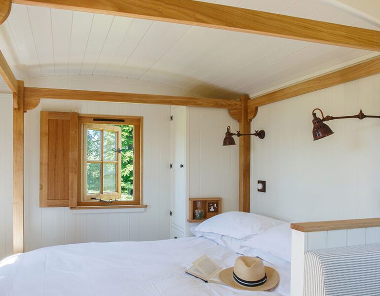 Bedroom with white walls, oak wood beams, a double bed, open window and a straw hat resting on the duvet.