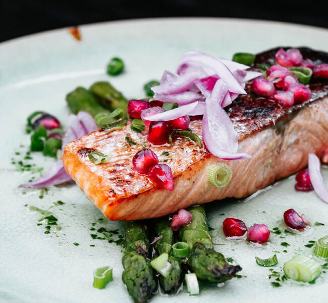 Seared salmon fillet served on asparagus, topped with red onion, pomegranate seeds, and chopped spring onions.