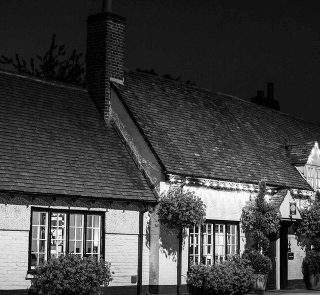 Black and white image of a traditional English pub with hanging flower baskets and a tiled roof at night.