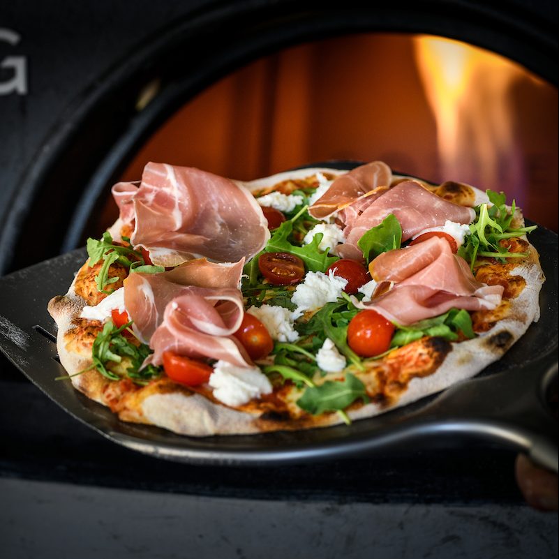 Wood-fired pizza topped with prosciutto, cherry tomatoes, rocket and mozzarella on a peel in front of a flame-lit oven.
