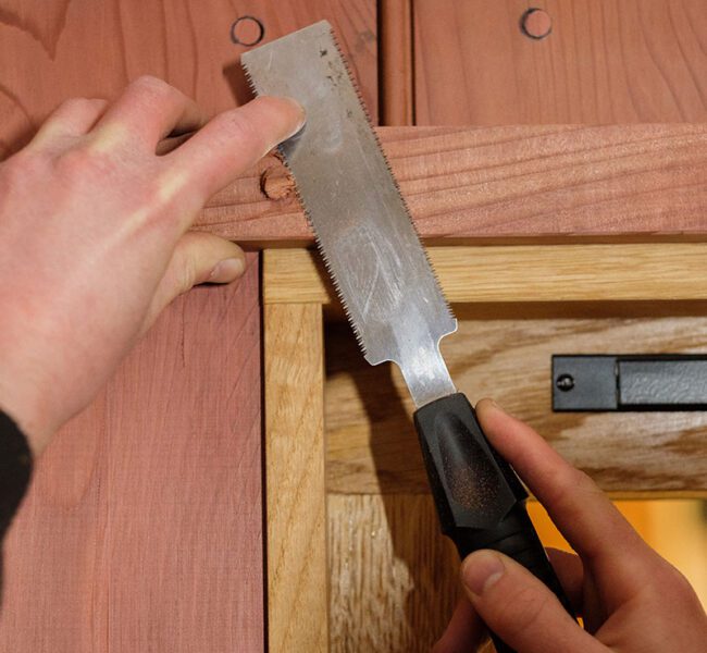 Close-up of hands using a fine-toothed saw to trim wood on a window frame set in red wooden panelling.