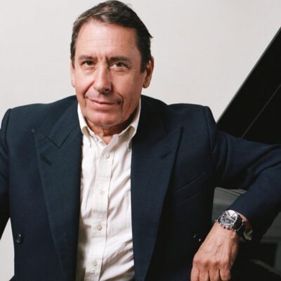 Musician Jools Holland in dark blazer and white shirt seated beside piano, looking at camera with relaxed expression.