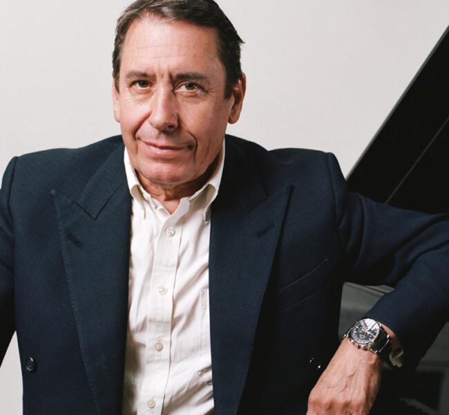 Musician Jools Holland in dark blazer and white shirt seated beside piano, looking at camera with relaxed expression.