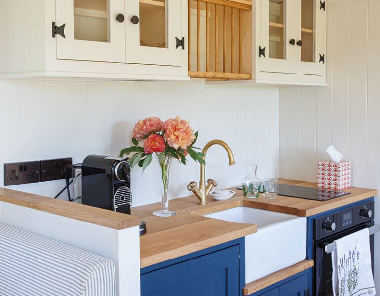 Small kitchen with navy and white units, farmhouse sink, brass tap, flowers on counter, cream wall cabinets with glass panels.