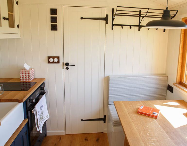 Compact kitchen and dining area in small cabin, with table, bench seating stove, farmhouse sink and white panelled walls.