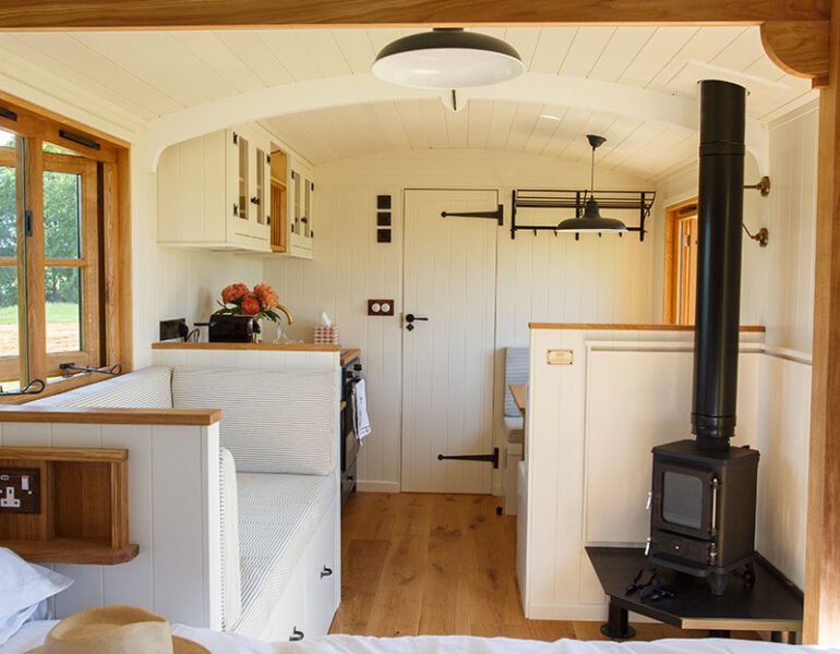 Bright drovers' hut interior with bed, kitchen, wood-burning stove, cushioned bench and dining area near the door.