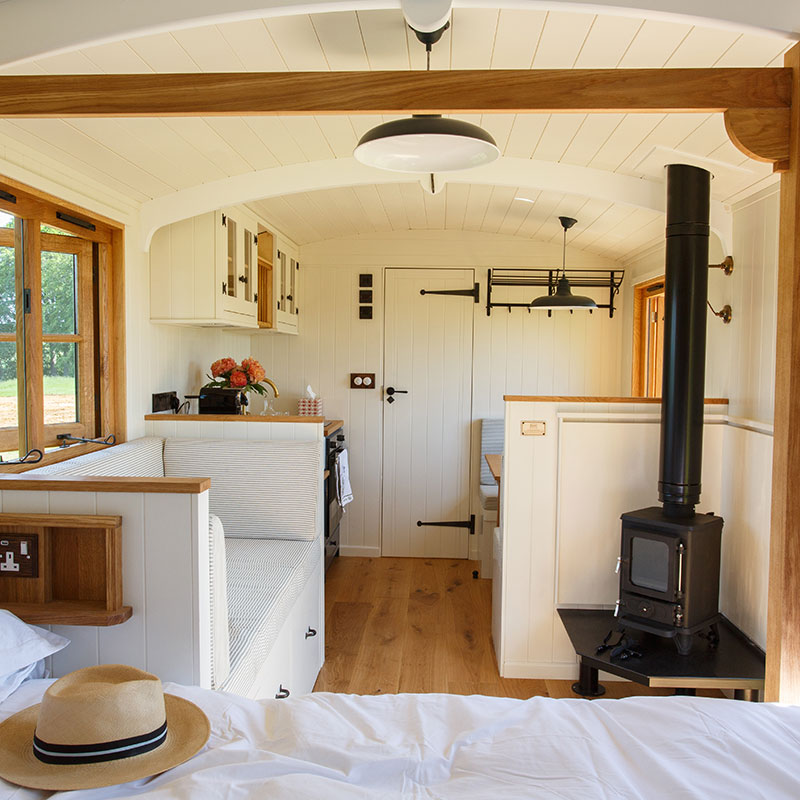 Bright drovers' hut interior with bed, kitchen, wood-burning stove, cushioned bench and dining area near the door.