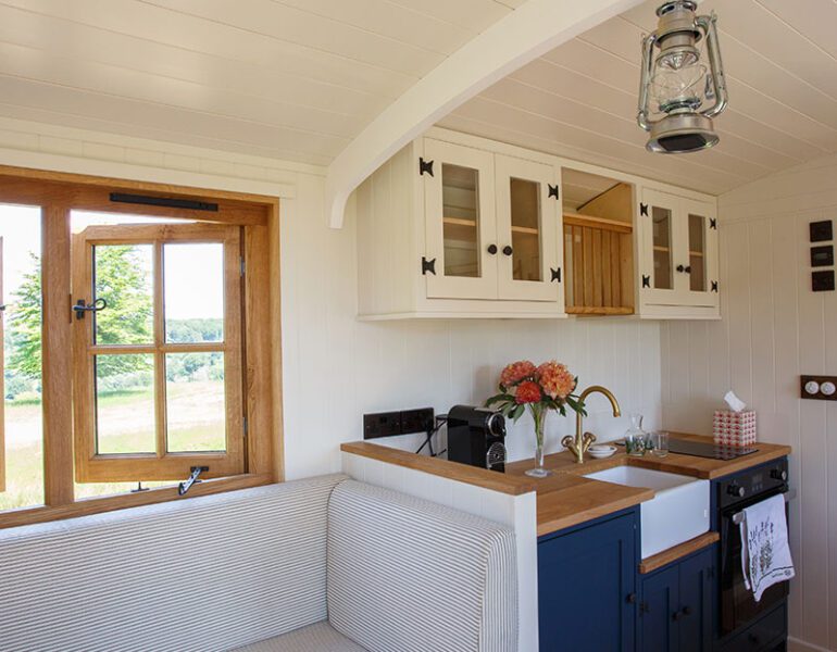 Drovers' hut kitchen with navy cabinets, white sink, cushioned bench, wooden windows and hanging lantern light.