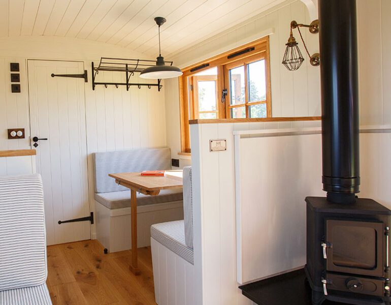 Compact wooden interior with seating nook, table, stove and light wood flooring in a small, cosy cabin or hut.