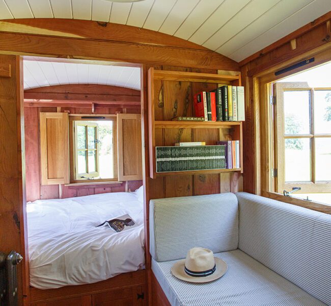 Drovers hut interior, cushioned window bench, bookshelves, window with field view and bedroom with bed in view.