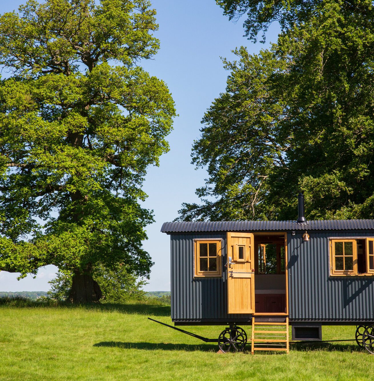 Black tin shepherd’s hut, wooden windows and door on wheels, set in a sunny green field with large trees in the background.