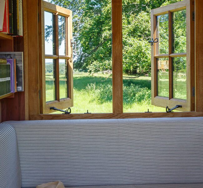 Open wooden-framed window in a cosy room with striped seating, looking out onto a sunny meadow and leafy trees.