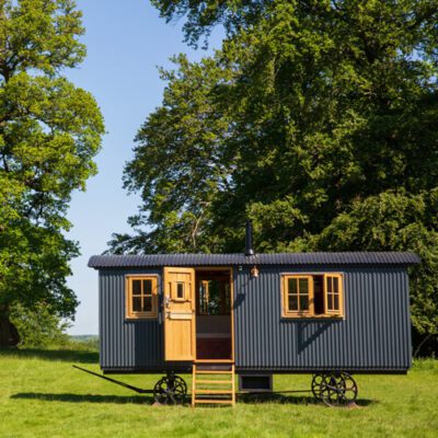 Black tin drovers’ hut, wooden windows and door on wheels, set in field with large trees in the background.