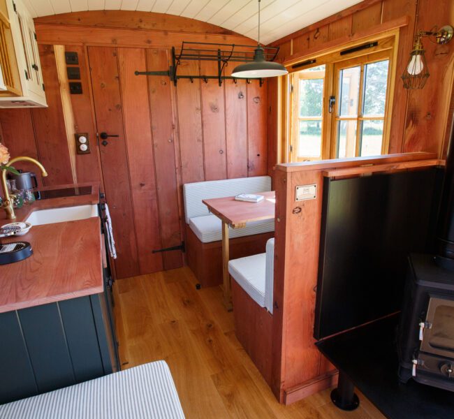 Interior of redwood drovers hut cabin with compact kitchen, dining booth by window and a black wood-burning stove.