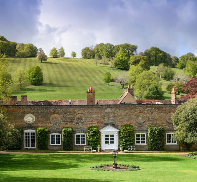 Large brick house with white door and ivy-clad walls, set before rolling green hills and trees under a dramatic sky.