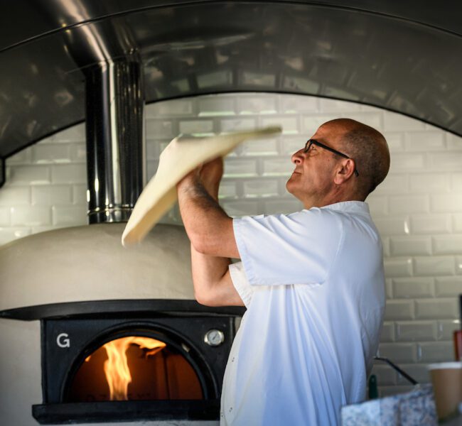 Chef tossing pizza dough in front of a wood-fired oven with white tiled walls in the background.