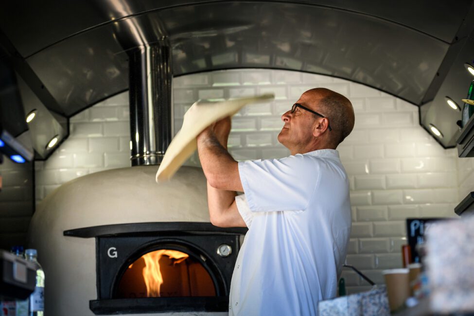 Chef tossing pizza dough in front of a wood-fired oven with white tiled walls in the background.
