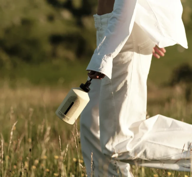 A person wearing white trousers and shirt walking through sunlit meadow, carrying a bottle of Commune body wash.