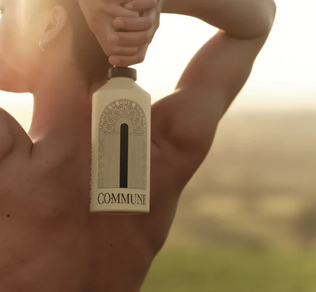 Person holding Commune body wash bottle behind their back, warm sunlight with blurred natural landscape in background.