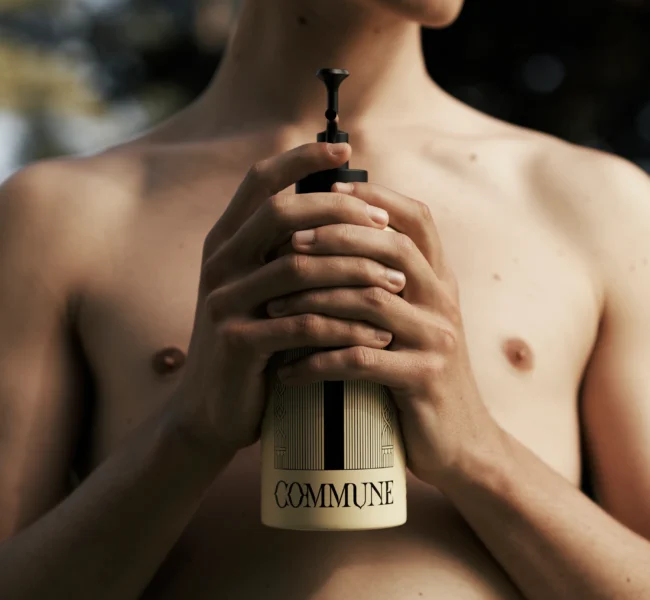 Shirtless man holding Commune body wash bottle in front of his body. Trees in background.